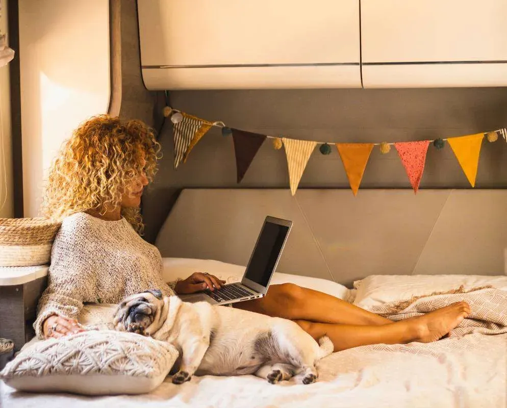 A woman working on a laptop in a duvet den strung with bunting, a pug asleep against her legs.