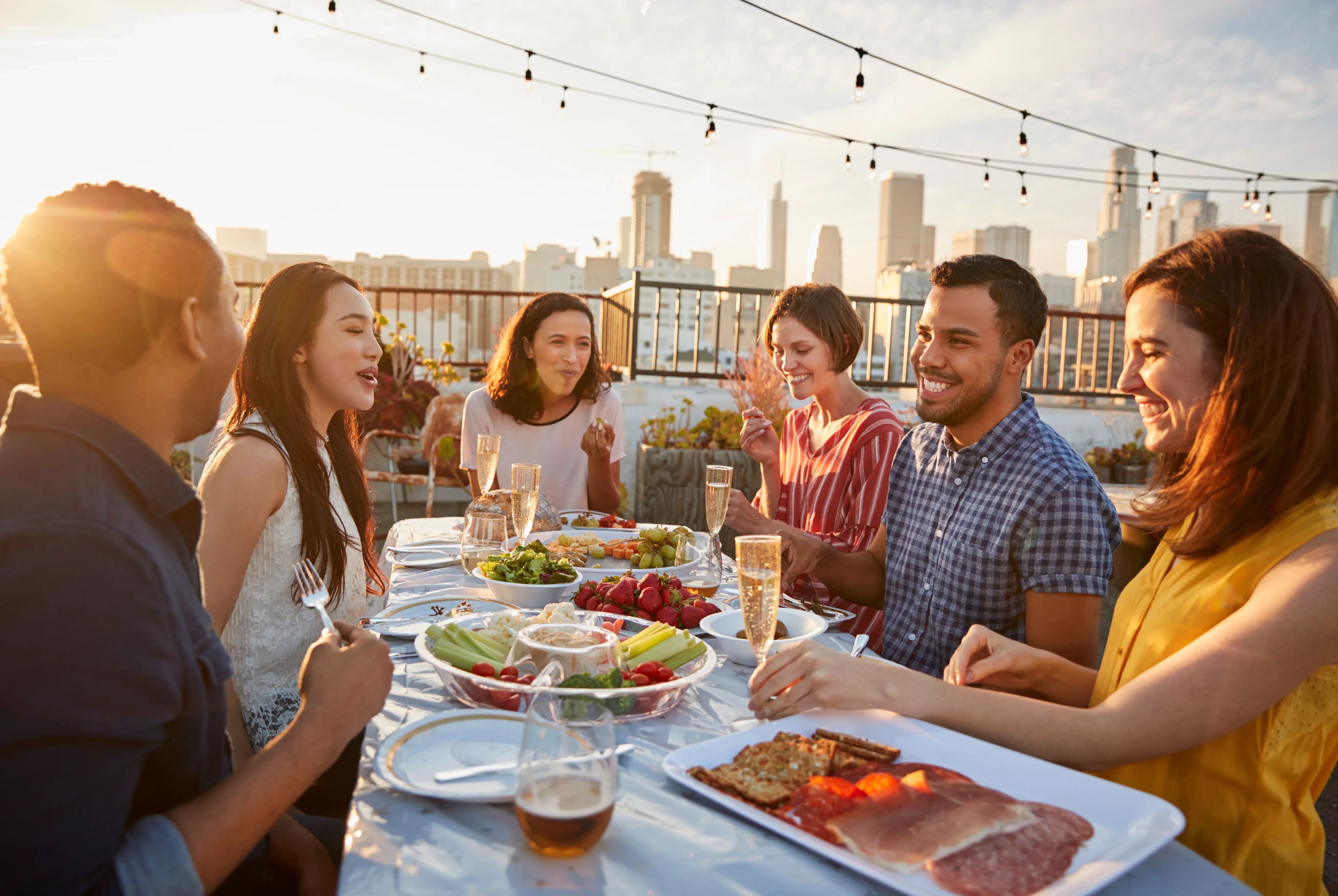 Friends sharing a meal around a long table on a rooftop terrace at dusk, festoon lights strung above and a city skyline behind.