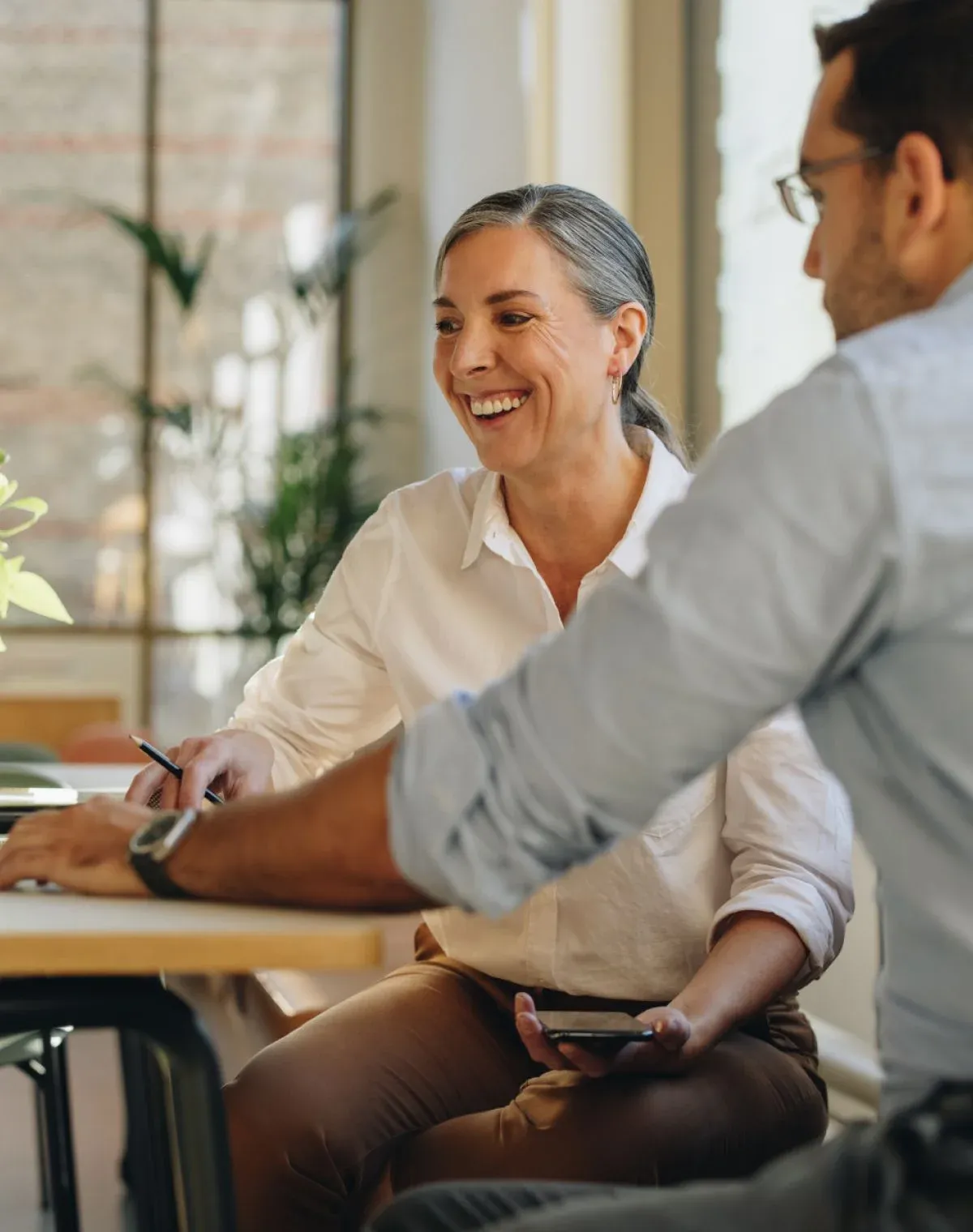 Two colleagues laughing together over a notebook at a table in a bright, plant-filled office.