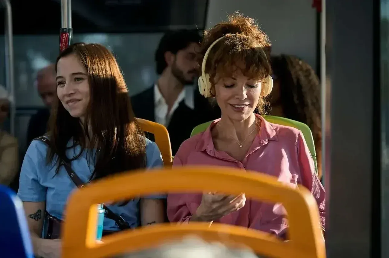 Two students sitting together on a bus, one wearing headphones, laughing as they talk.