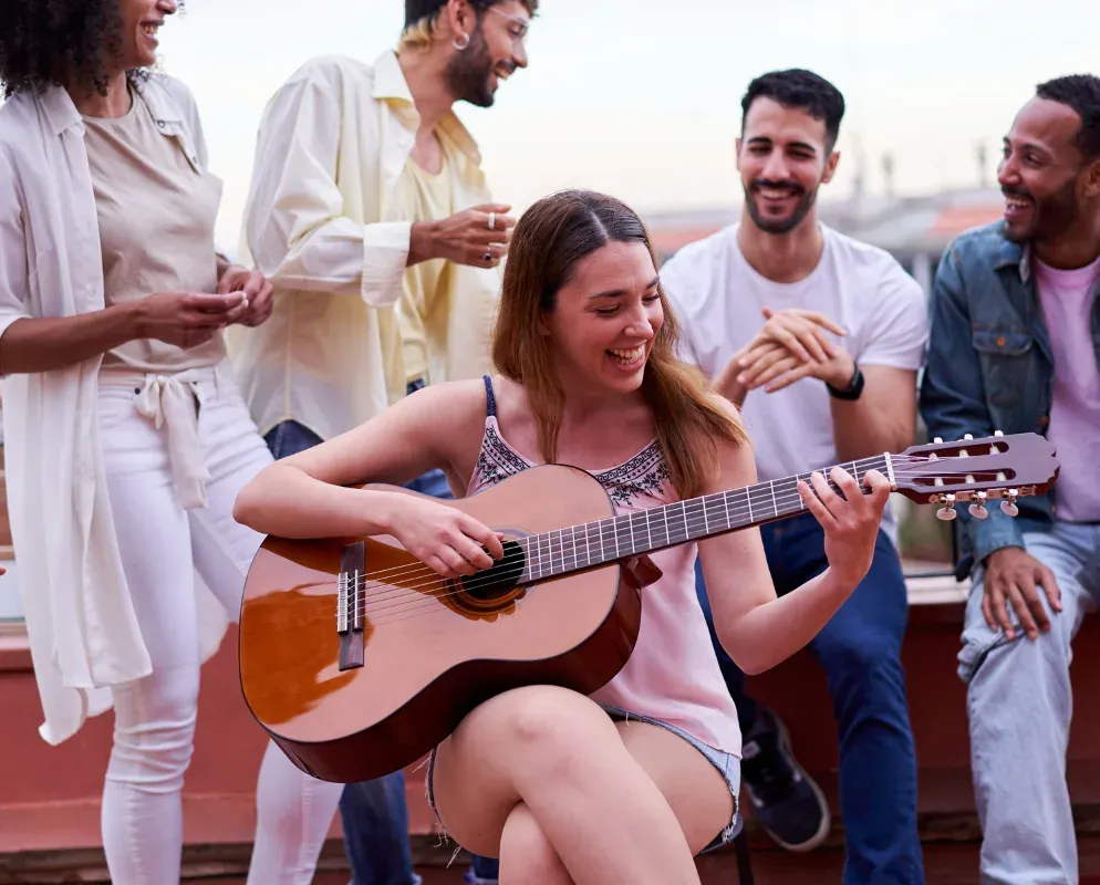 A group of friends on a rooftop at dusk, one playing an acoustic guitar while the others listen.