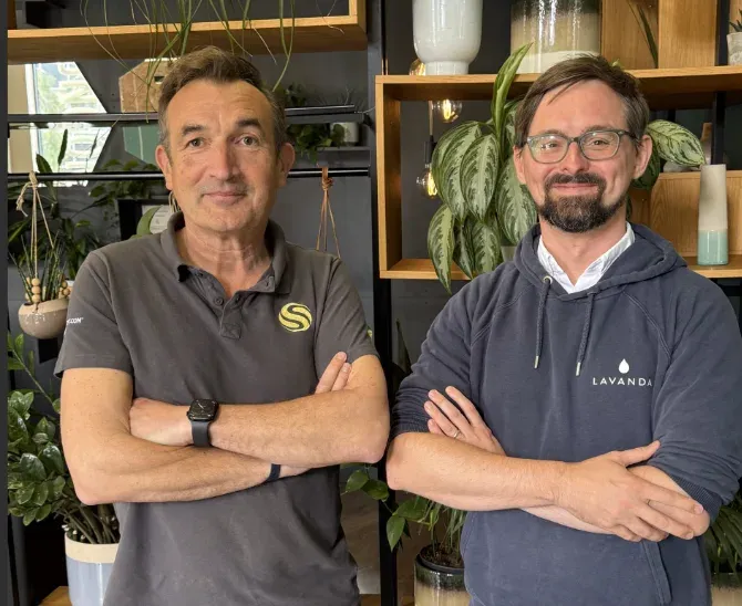 Two men standing side by side among houseplants, one in a Student.com polo shirt and one in a Lavanda hoodie.