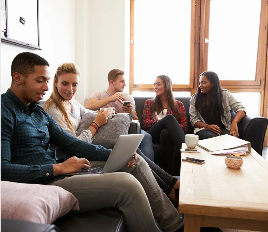 Five students sitting with laptops and mugs in the daylit lounge of a shared house.