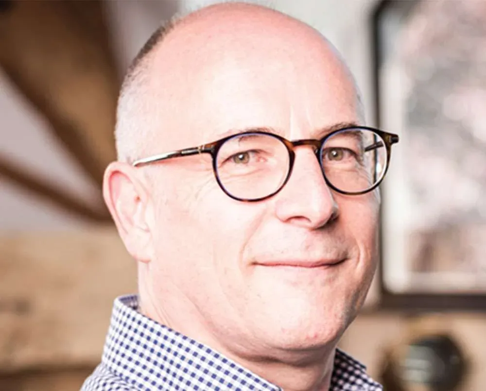 A headshot of a man in dark-rimmed glasses and a checked shirt.